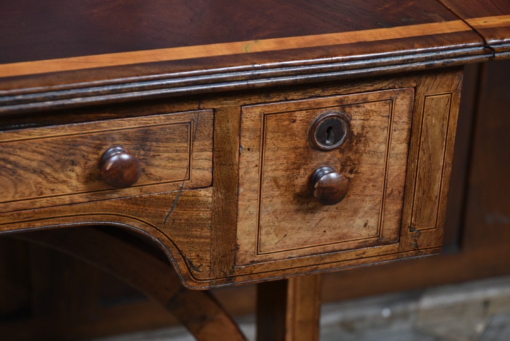 A Regency rosewood sofa table featuring a crossbanded drop-leaf top, shallow drawers with wooden knobs, curved trestle legs, and brass castors. View VI