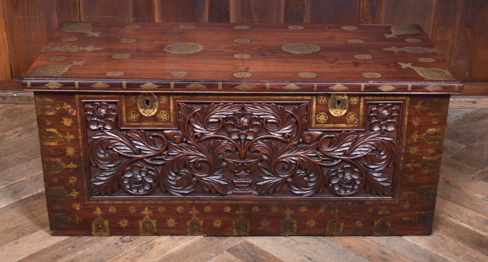 A dark reddish brown padouk wood storage chest featuring a relief-carved floral front panel and decorative brass metal mounts across the top lid and frame. View II