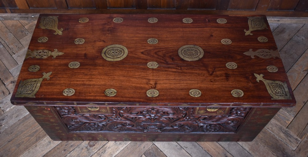 A dark reddish brown padouk wood storage chest featuring a relief-carved floral front panel and decorative brass metal mounts across the top lid and frame. View V