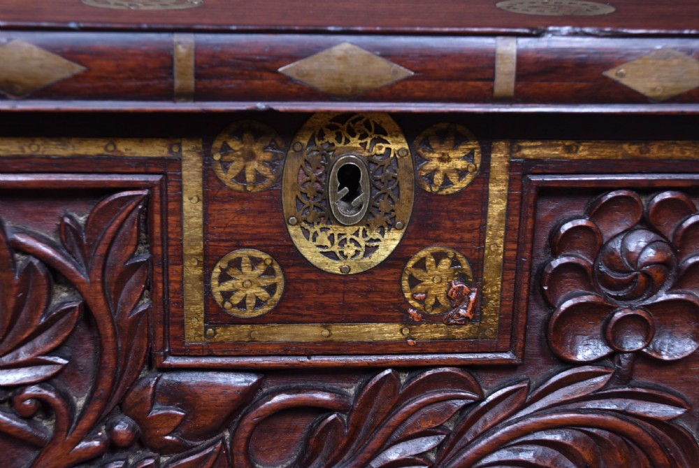 A dark reddish brown padouk wood storage chest featuring a relief-carved floral front panel and decorative brass metal mounts across the top lid and frame. View VI