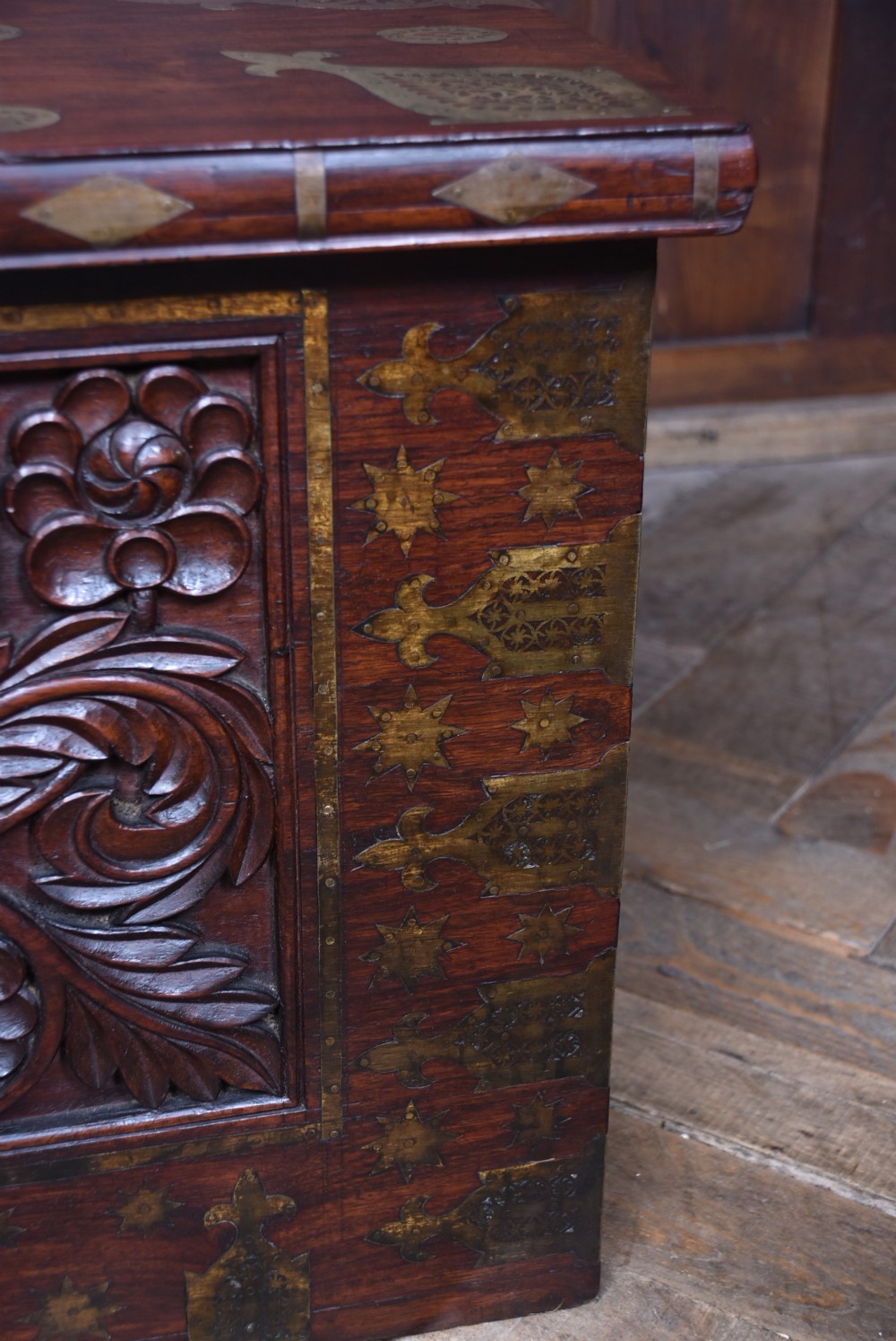 A dark reddish brown padouk wood storage chest featuring a relief-carved floral front panel and decorative brass metal mounts across the top lid and frame. View VII