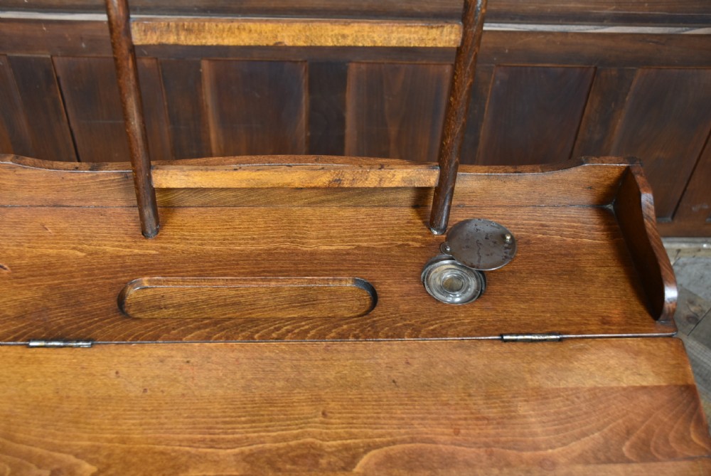 An antique Edwardian oak school desk featuring a slanted writing top, an attached slatted seat, lower shelf slats, and geometric marquetry inlay on the side panel. View XV
