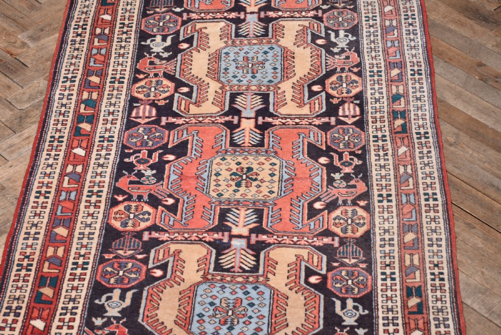 A hand-knotted wool runner rug featuring geometric medallions, bird motifs, and decorative borders in terracotta, cream, blue, and dark navy tones. View III
