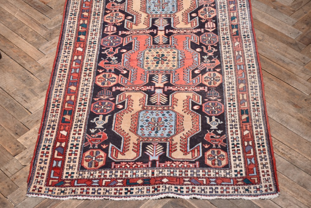 A hand-knotted wool runner rug featuring geometric medallions, bird motifs, and decorative borders in terracotta, cream, blue, and dark navy tones. View V