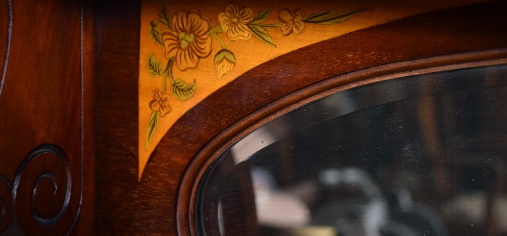 An Edwardian mahogany display cabinet featuring hand-painted floral borders, an oval mirror top section, glazed doors, and cabriole legs. View X