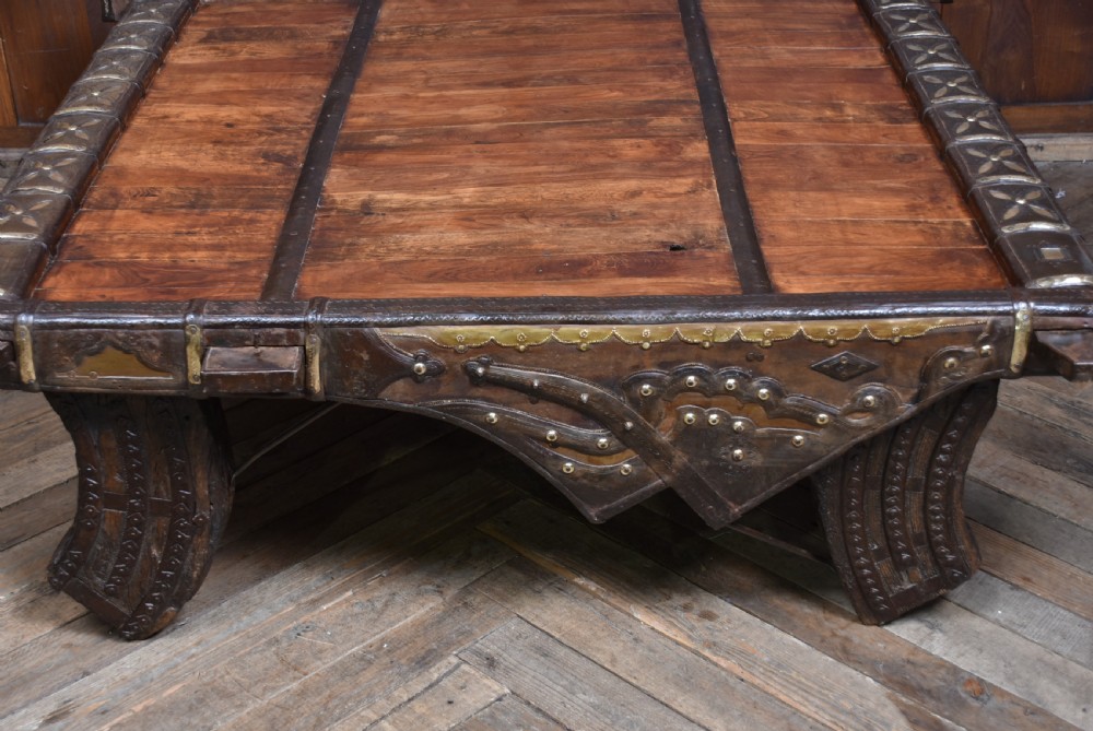A low square coffee table crafted from a dark reclaimed bullock cart frame with decorative brass studding and stout carved legs. View VI