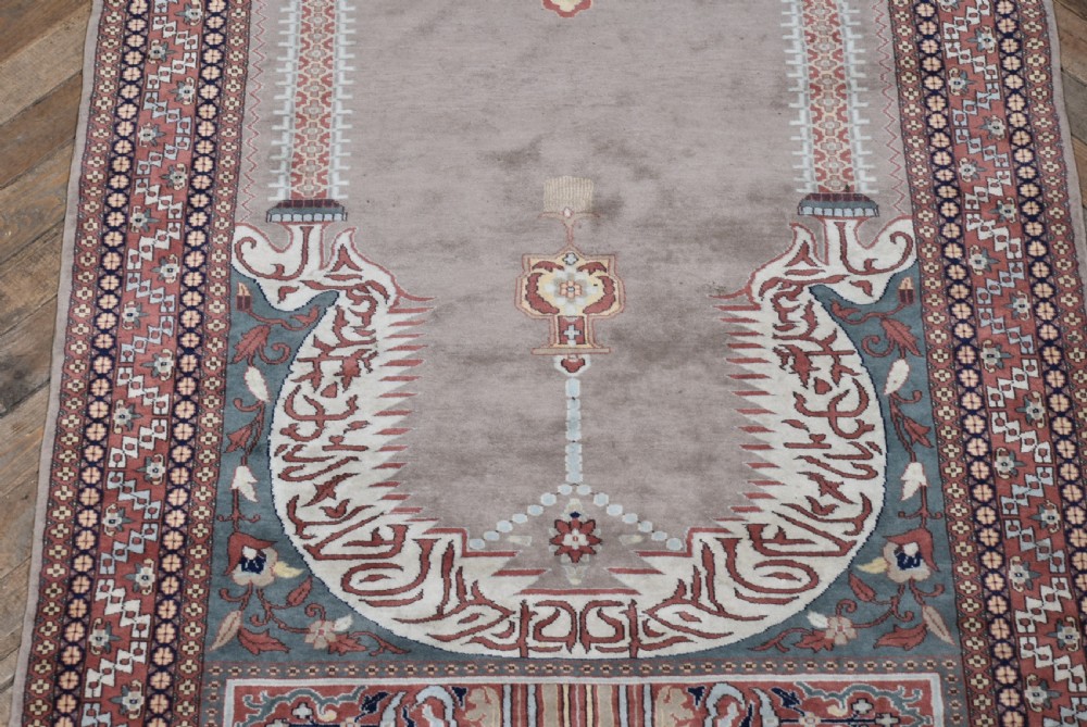 A hand-woven silk prayer rug featuring a central arched mihrab motif in taupe, framed by intricate terracotta and slate blue geometric floral borders with fringe at both ends. View V