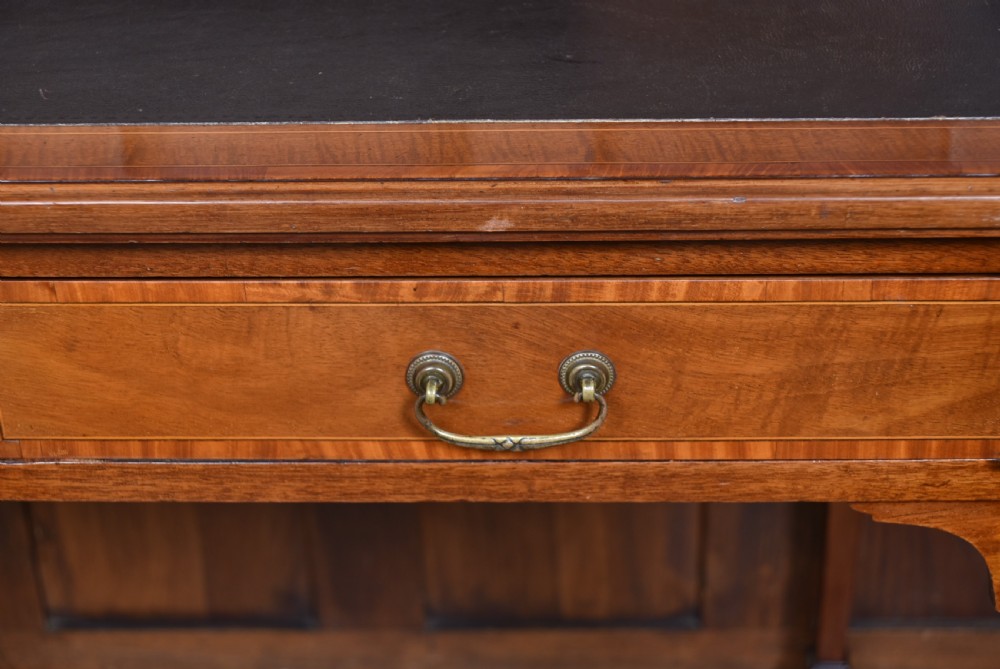 An Edwardian mahogany writing desk featuring a leather writing surface, inlaid drawer fronts, a brass gallery, and slim tapered legs on castors. View V