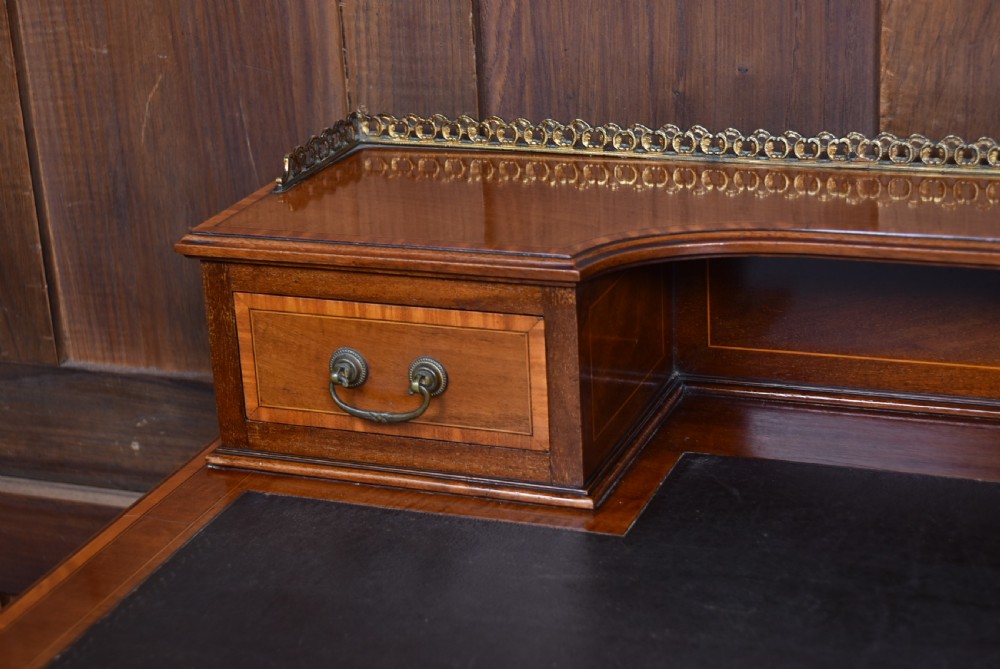 An Edwardian mahogany writing desk featuring a leather writing surface, inlaid drawer fronts, a brass gallery, and slim tapered legs on castors. View VI