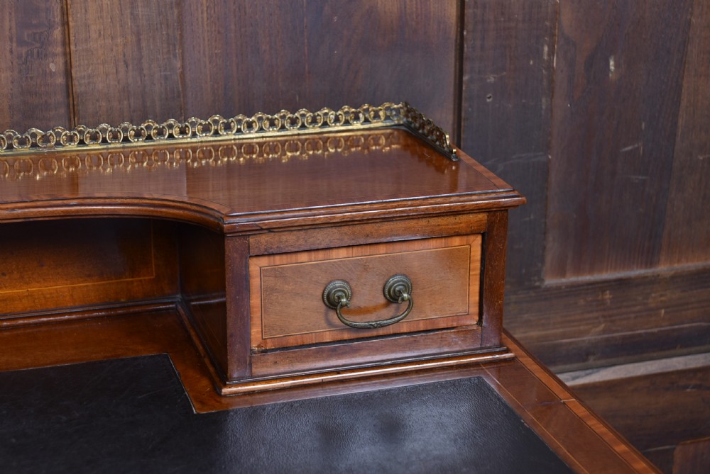 An Edwardian mahogany writing desk featuring a leather writing surface, inlaid drawer fronts, a brass gallery, and slim tapered legs on castors. View VII