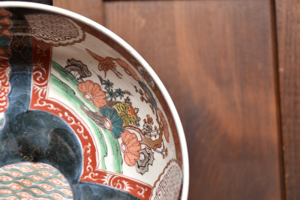 A round Japanese Imari porcelain bowl styled with cobalt blue and iron-red panelled decoration, resting on a carved dark wooden display stand. View III