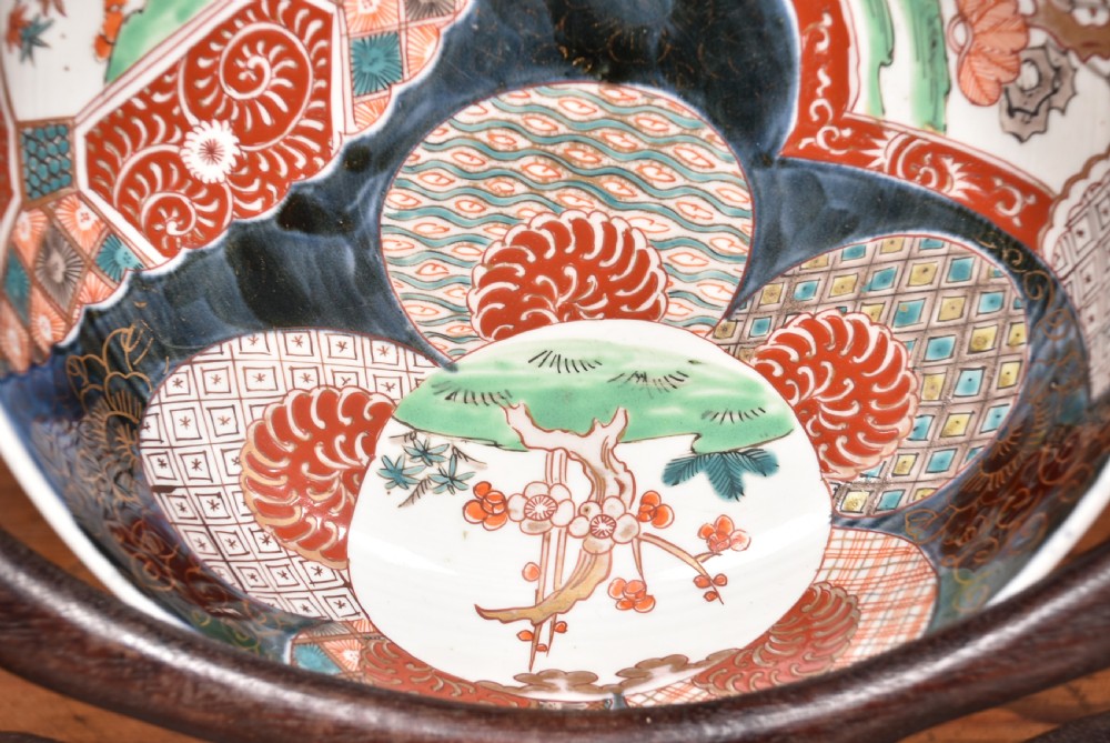 A round Japanese Imari porcelain bowl styled with cobalt blue and iron-red panelled decoration, resting on a carved dark wooden display stand. View V