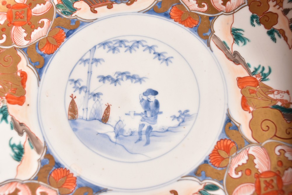 A circular Japanese Imari porcelain bowl decorated with red, blue, green, and gold floral patterns, displayed on a dark carved wood stand. View III