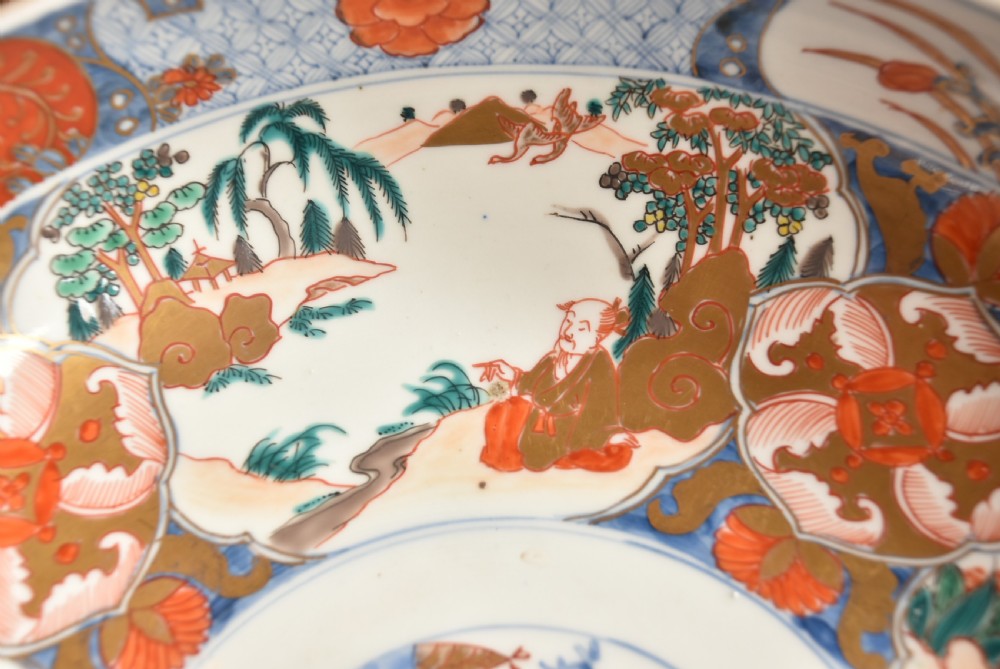 A circular Japanese Imari porcelain bowl decorated with red, blue, green, and gold floral patterns, displayed on a dark carved wood stand. View IV