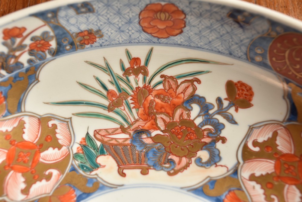 A circular Japanese Imari porcelain bowl decorated with red, blue, green, and gold floral patterns, displayed on a dark carved wood stand. View V