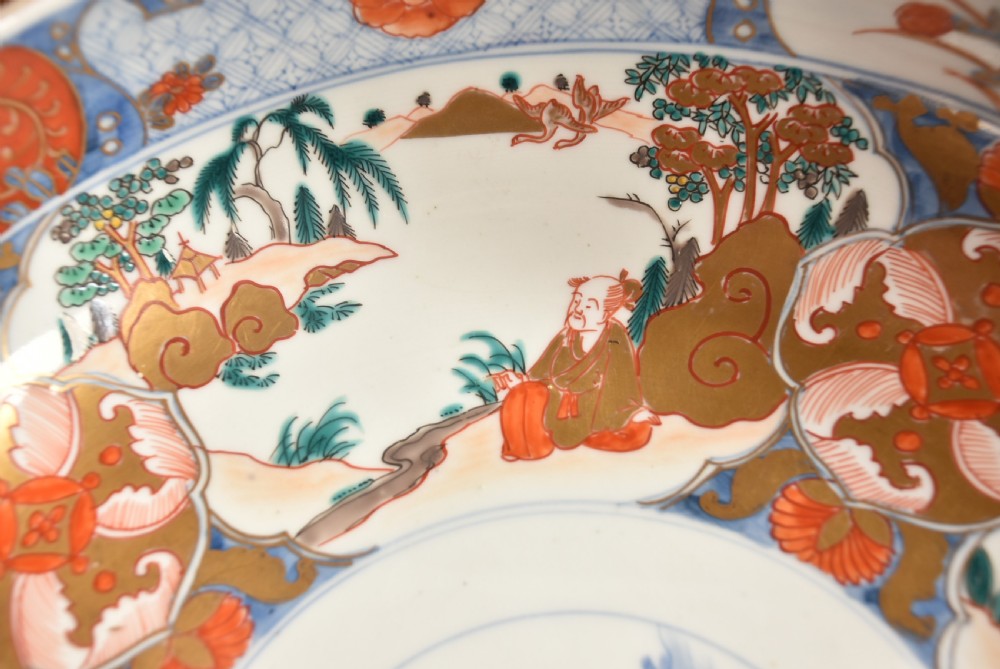 A circular Japanese Imari porcelain bowl decorated with red, blue, green, and gold floral patterns, displayed on a dark carved wood stand. View VI