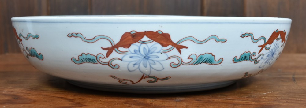 A circular Japanese Imari porcelain bowl decorated with red, blue, green, and gold floral patterns, displayed on a dark carved wood stand. View VII