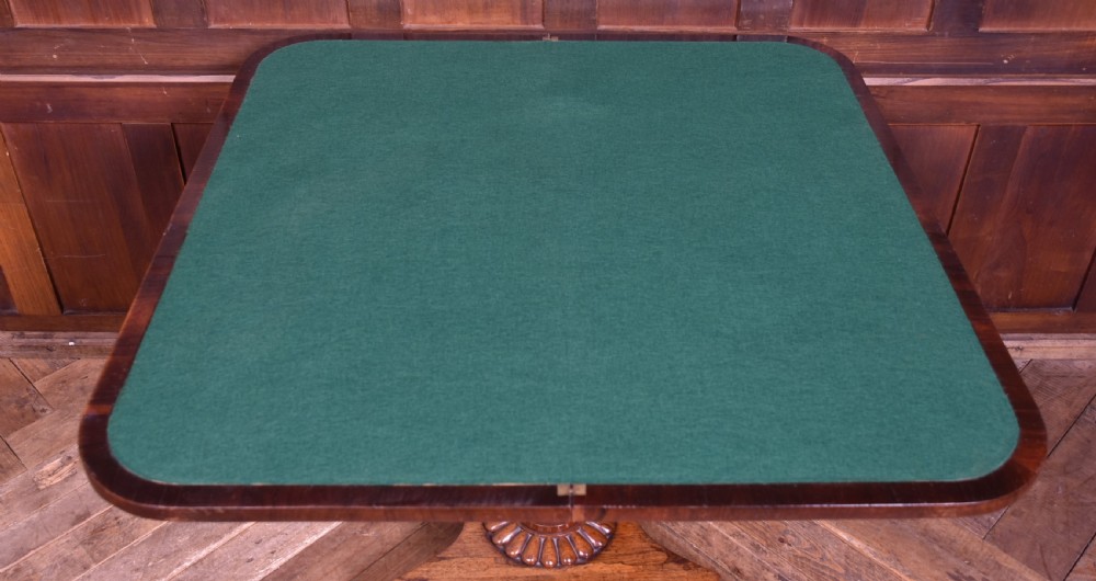 A Victorian rosewood fold-over games table featuring a polished rectangular top, turned carved pedestal stem, and a quadripartite plinth base with carved claw feet. View X