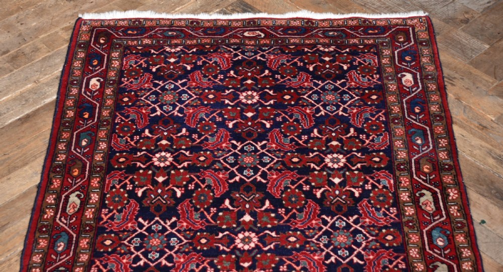 A rectangular hand-knotted wool Persian rug featuring a dark navy blue central field filled with intricate red floral lattice patterns and surrounded by decorative floral borders with short end fringe. View III