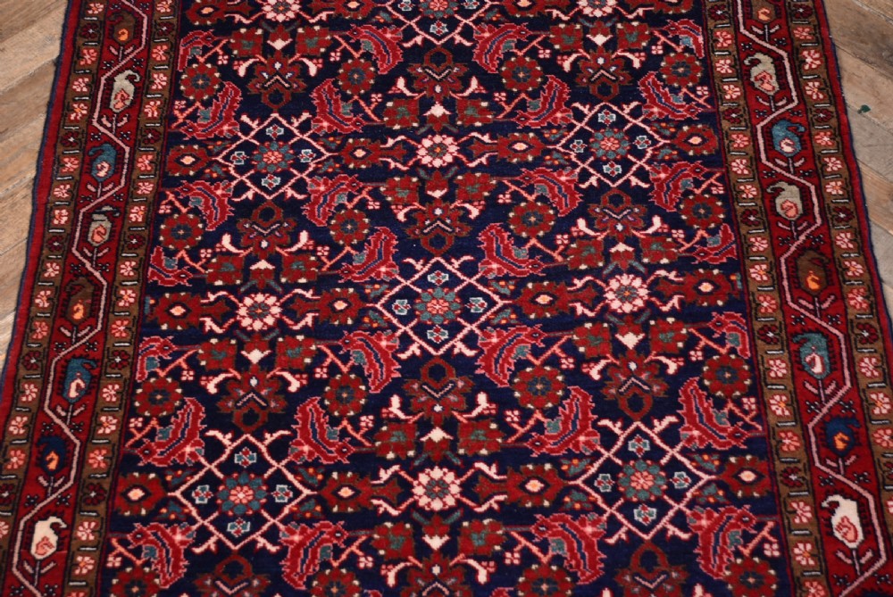 A rectangular hand-knotted wool Persian rug featuring a dark navy blue central field filled with intricate red floral lattice patterns and surrounded by decorative floral borders with short end fringe. View V