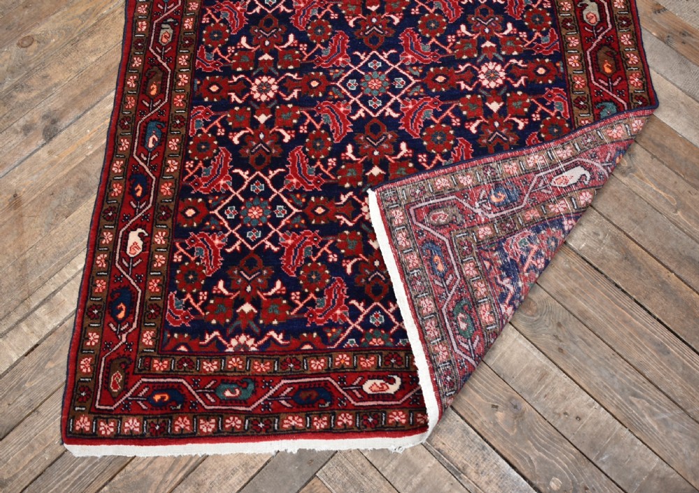 A rectangular hand-knotted wool Persian rug featuring a dark navy blue central field filled with intricate red floral lattice patterns and surrounded by decorative floral borders with short end fringe. View VII