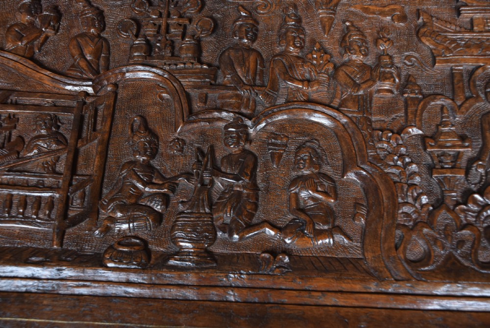 A 19th-century dark teak storage chest featuring detailed relief carving of figures and architecture on the front and side panels, complete with brass latches and corner straps. View V