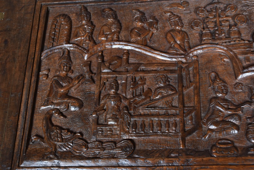 A 19th-century dark teak storage chest featuring detailed relief carving of figures and architecture on the front and side panels, complete with brass latches and corner straps. View VI