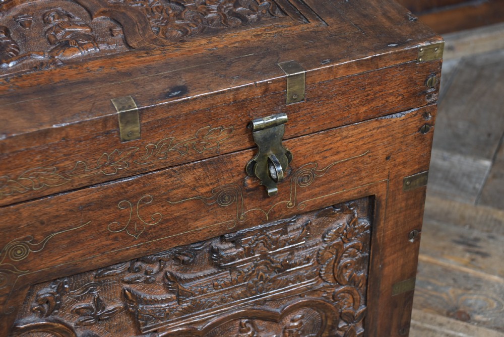 A 19th-century dark teak storage chest featuring detailed relief carving of figures and architecture on the front and side panels, complete with brass latches and corner straps. View VIII