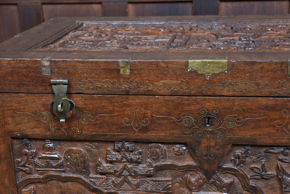 A 19th-century dark teak storage chest featuring detailed relief carving of figures and architecture on the front and side panels, complete with brass latches and corner straps. View IX
