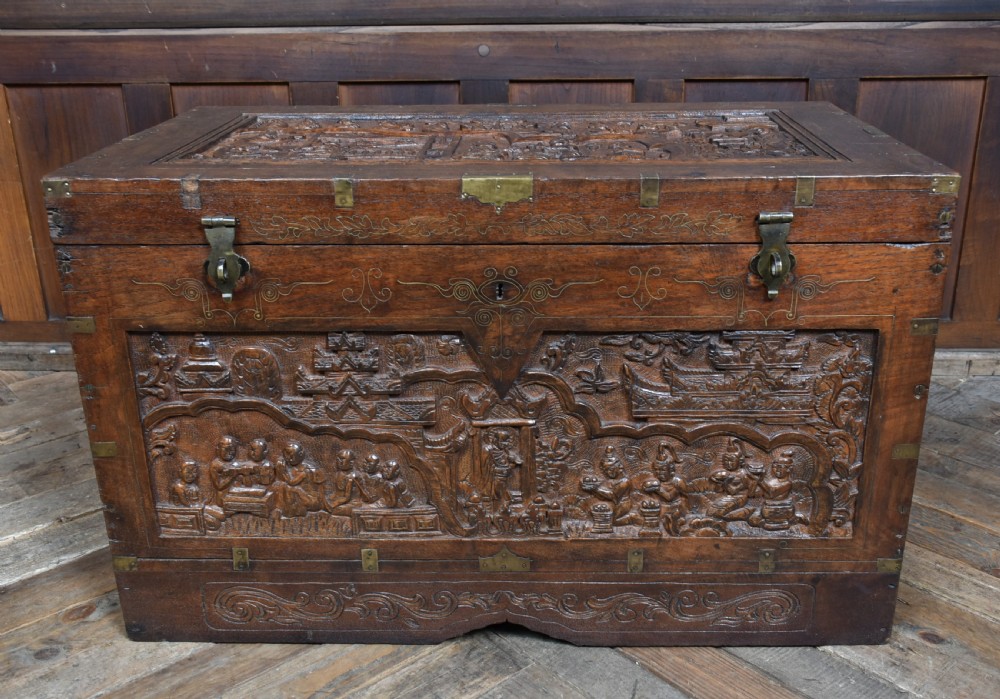 A 19th-century dark teak storage chest featuring detailed relief carving of figures and architecture on the front and side panels, complete with brass latches and corner straps. View X
