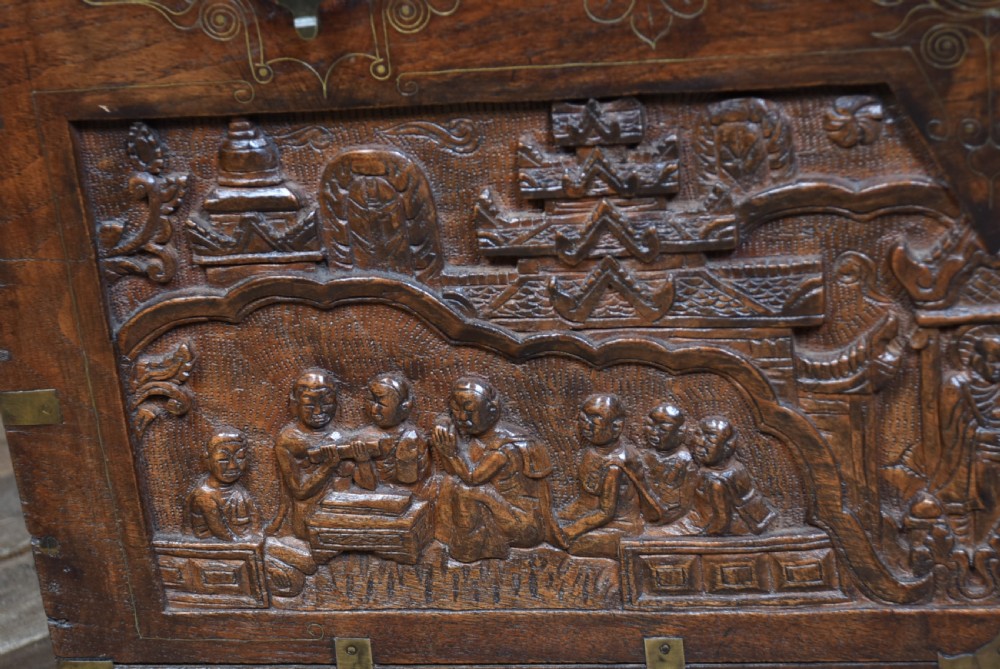 A 19th-century dark teak storage chest featuring detailed relief carving of figures and architecture on the front and side panels, complete with brass latches and corner straps. View XI