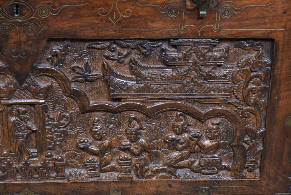 A 19th-century dark teak storage chest featuring detailed relief carving of figures and architecture on the front and side panels, complete with brass latches and corner straps. View XII