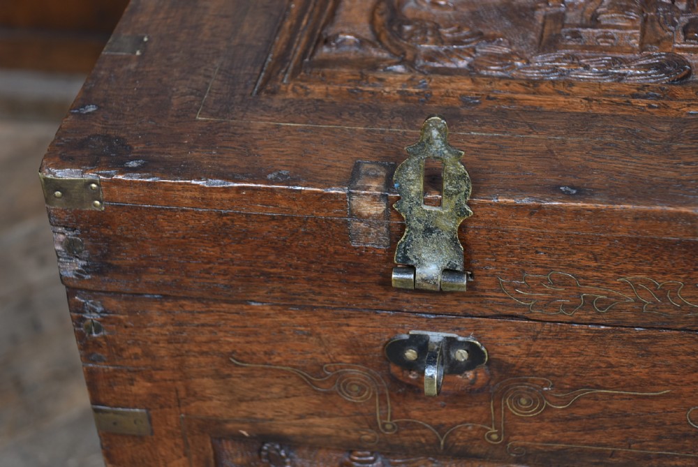 A 19th-century dark teak storage chest featuring detailed relief carving of figures and architecture on the front and side panels, complete with brass latches and corner straps. View XIII
