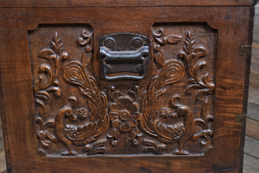 A 19th-century dark teak storage chest featuring detailed relief carving of figures and architecture on the front and side panels, complete with brass latches and corner straps. View XIX