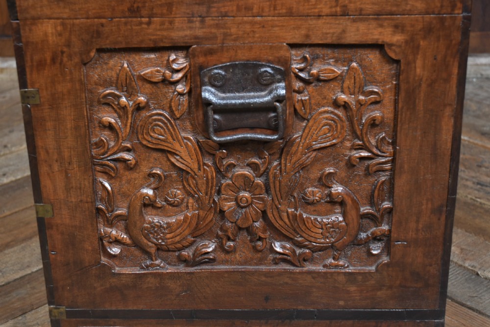 A 19th-century dark teak storage chest featuring detailed relief carving of figures and architecture on the front and side panels, complete with brass latches and corner straps. View XXII