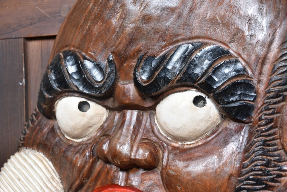 A hand-carved wooden wall plaque depicting Daruma with pronounced black eyebrows, wide white eyes, a wide moustache, and a red lacquered border. View IV