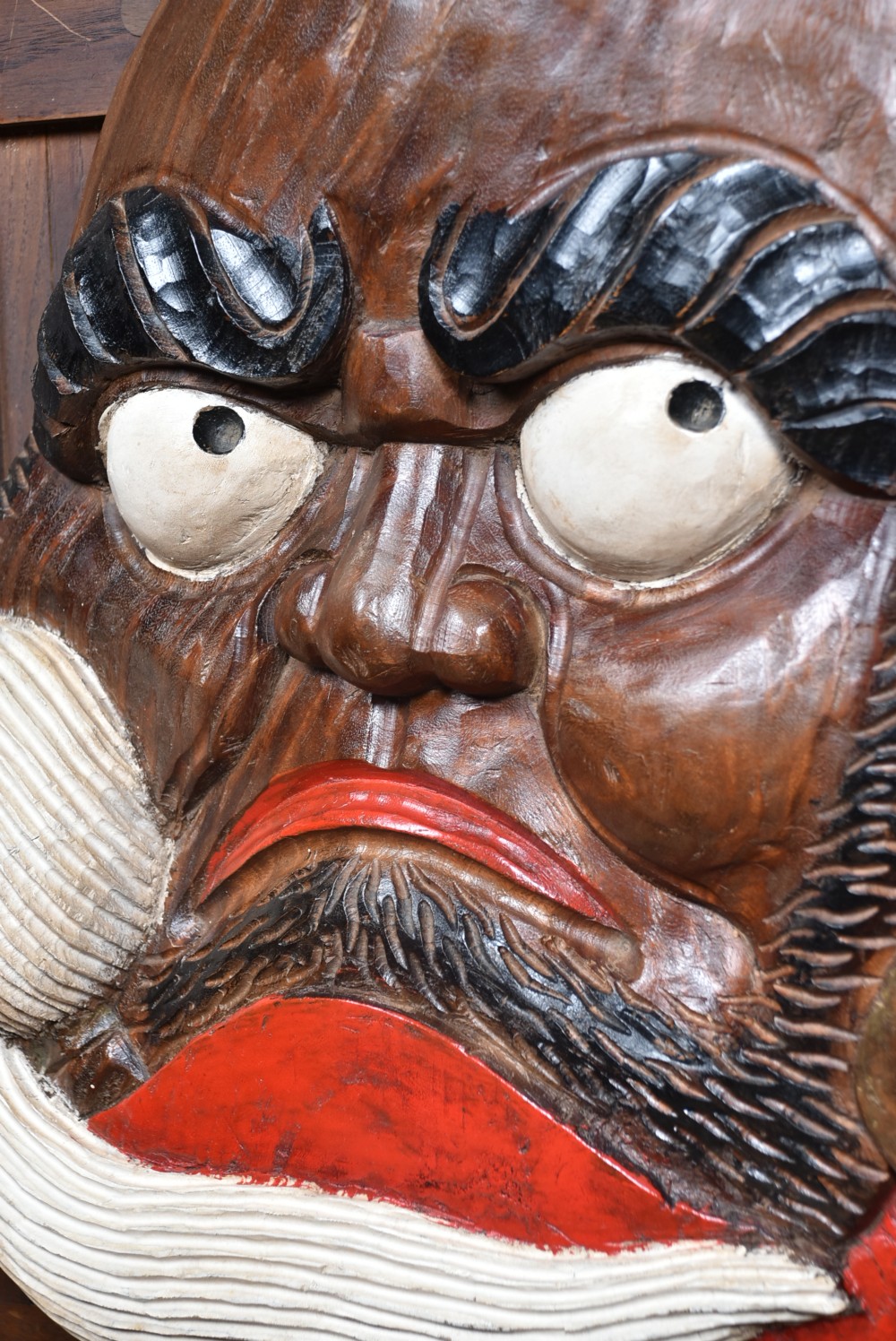 A hand-carved wooden wall plaque depicting Daruma with pronounced black eyebrows, wide white eyes, a wide moustache, and a red lacquered border. View V