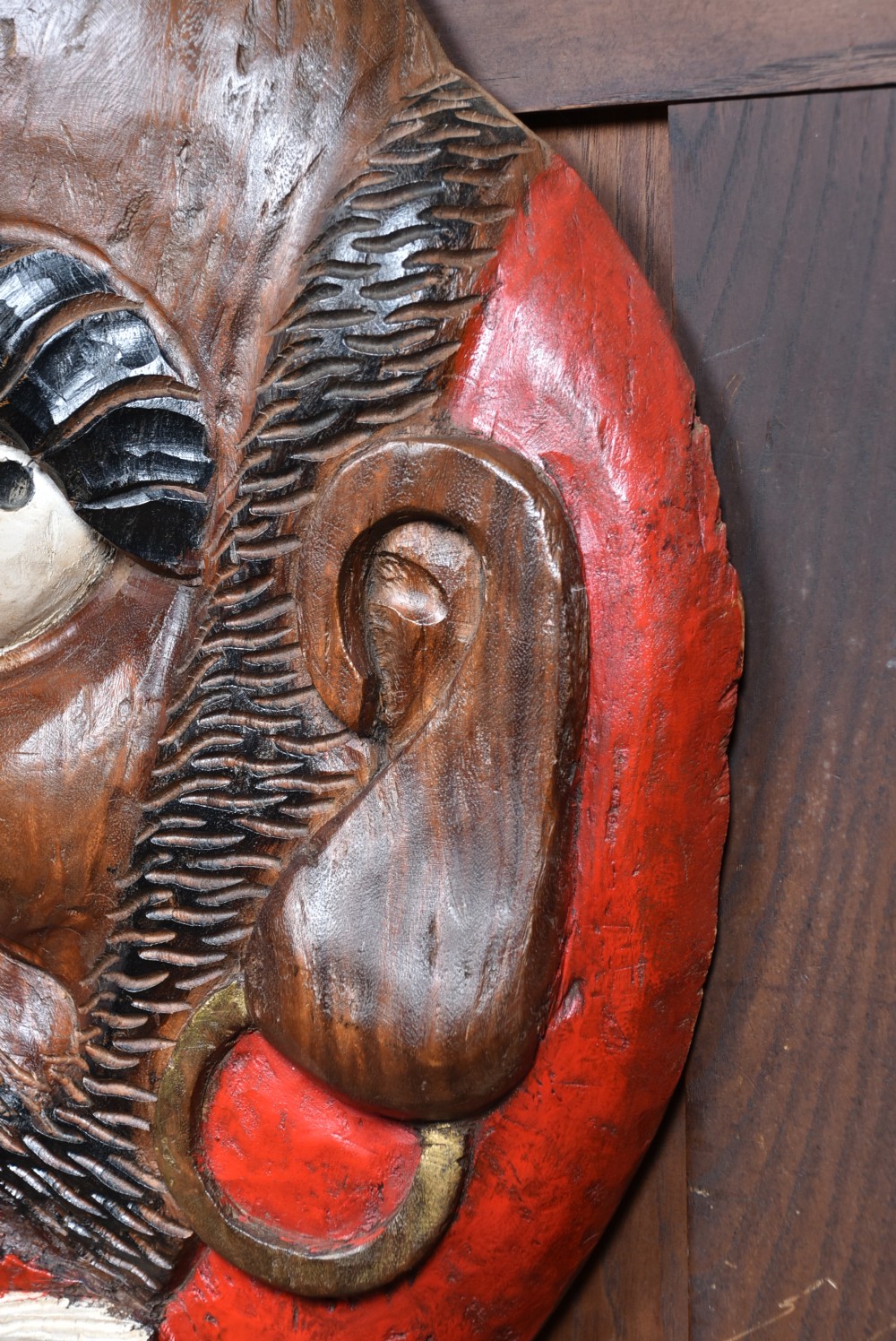 A hand-carved wooden wall plaque depicting Daruma with pronounced black eyebrows, wide white eyes, a wide moustache, and a red lacquered border. View VI
