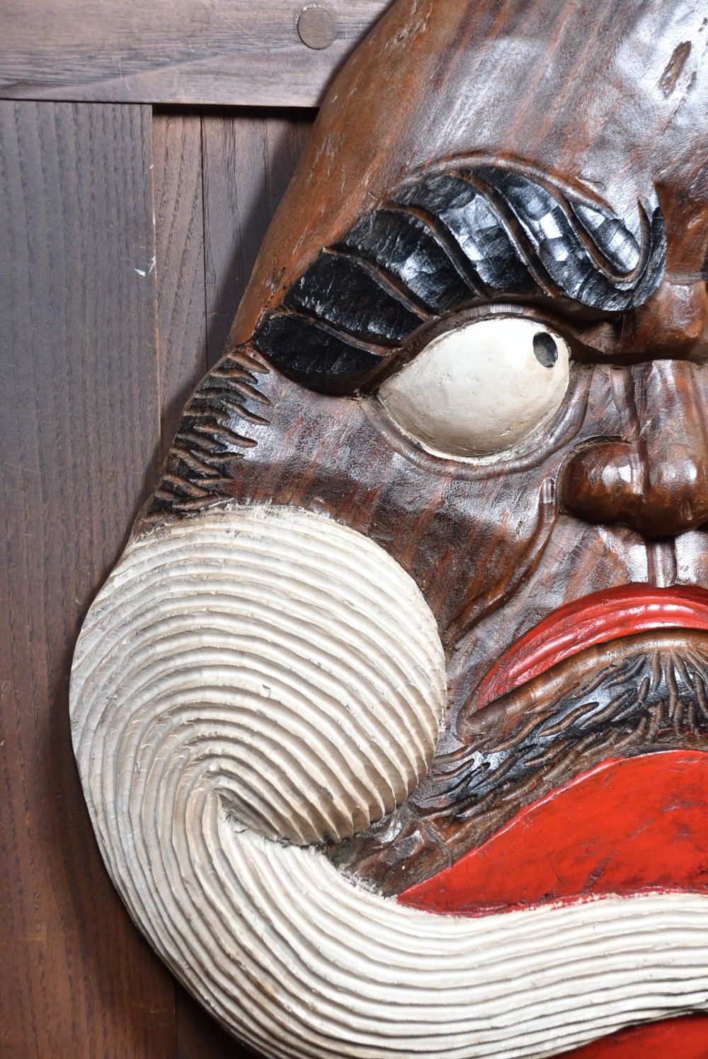 A hand-carved wooden wall plaque depicting Daruma with pronounced black eyebrows, wide white eyes, a wide moustache, and a red lacquered border. View VII