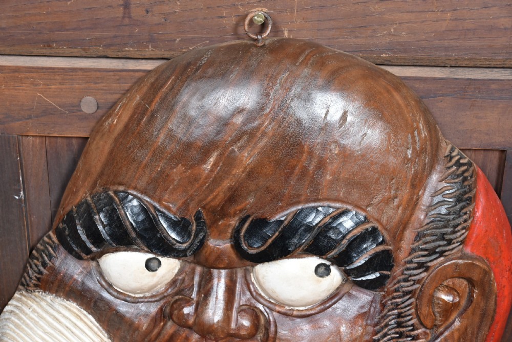 A hand-carved wooden wall plaque depicting Daruma with pronounced black eyebrows, wide white eyes, a wide moustache, and a red lacquered border. View VIII