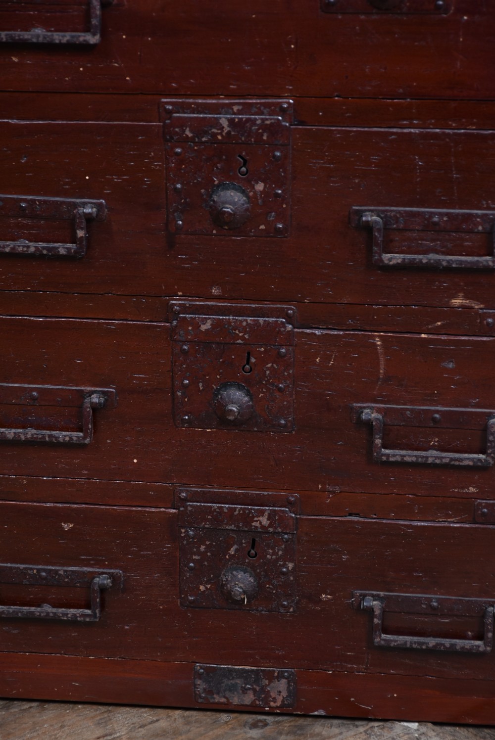 A Japanese dark red lacquered wood tansu chest featuring heavy iron corner mounts, bail handles, and multiple drawers of varying sizes. View VI