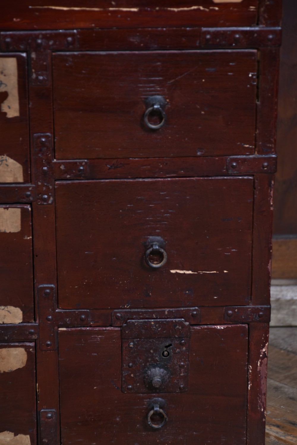 A Japanese dark red lacquered wood tansu chest featuring heavy iron corner mounts, bail handles, and multiple drawers of varying sizes. View VII