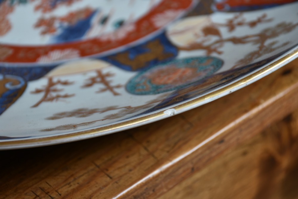 A decorated circular Japanese Imari porcelain charger with blue, red, and gold floral and landscape motifs, resting inside a square wooden storage box alongside its lid. View V