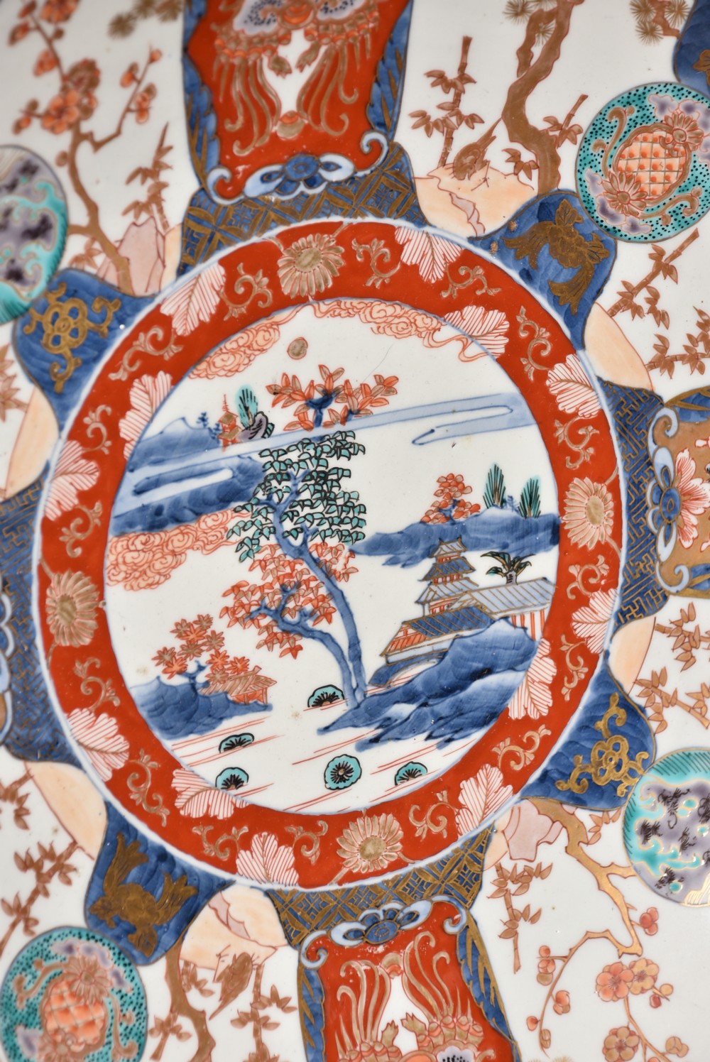 A decorated circular Japanese Imari porcelain charger with blue, red, and gold floral and landscape motifs, resting inside a square wooden storage box alongside its lid. View VII