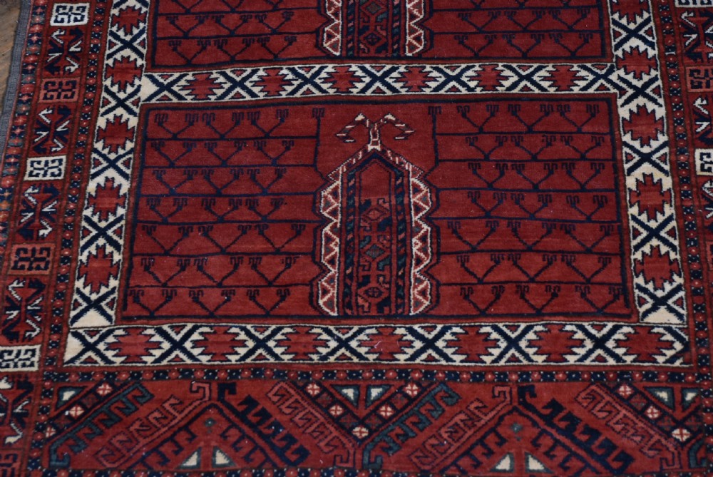 A handwoven red wool rug featuring double arched niche motifs, geometric border patterns, dark navy details, and fringe ends. View V