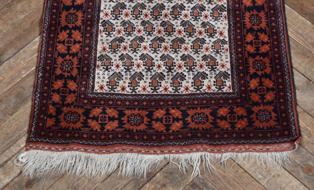 A hand-woven wool rug featuring a cream central field with repeating geometric motifs surrounded by a wide rust-red patterned border and white fringe. View III