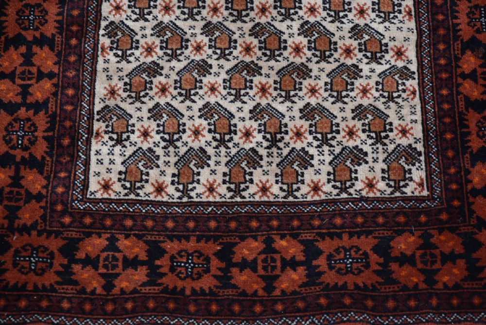 A hand-woven wool rug featuring a cream central field with repeating geometric motifs surrounded by a wide rust-red patterned border and white fringe. View V