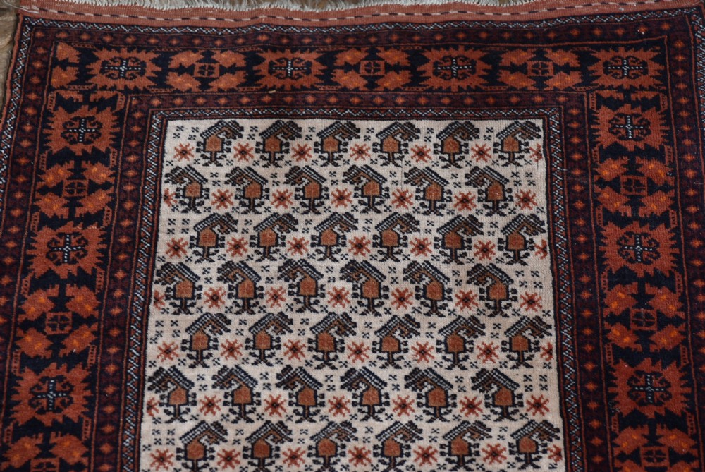 A hand-woven wool rug featuring a cream central field with repeating geometric motifs surrounded by a wide rust-red patterned border and white fringe. View VII