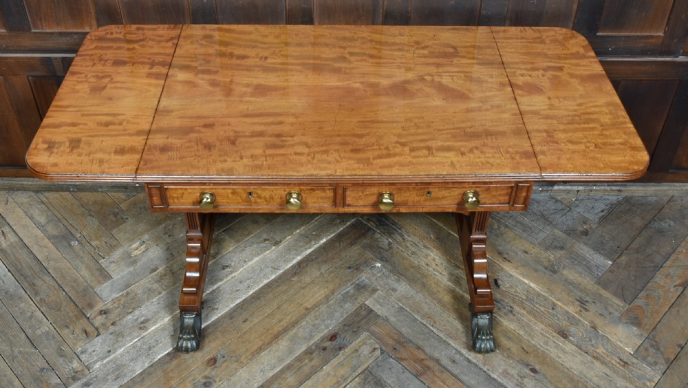 A Regency mahogany drop-leaf sofa table featuring flame mahogany drawer fronts, reeded upright trestle legs, a stretcher with central roundel, and brass lion paw castors. View X