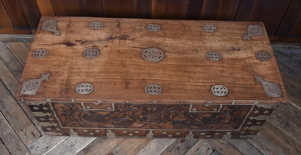A low rectangular carved wood storage chest featuring decorative brass mounts, corner brackets, studs, and a central relief carving of a floral motif. View III